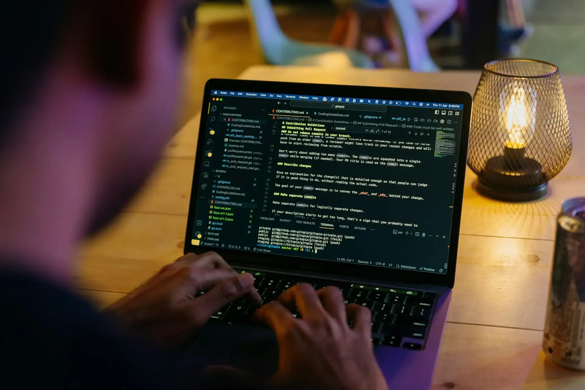 Widok z góry na ręce programisty piszące na klawiaturze laptopa, na ekranie otwarty edytor kodu z plikiem Markdown, w tle rozmyta lampka na drewnianym stole